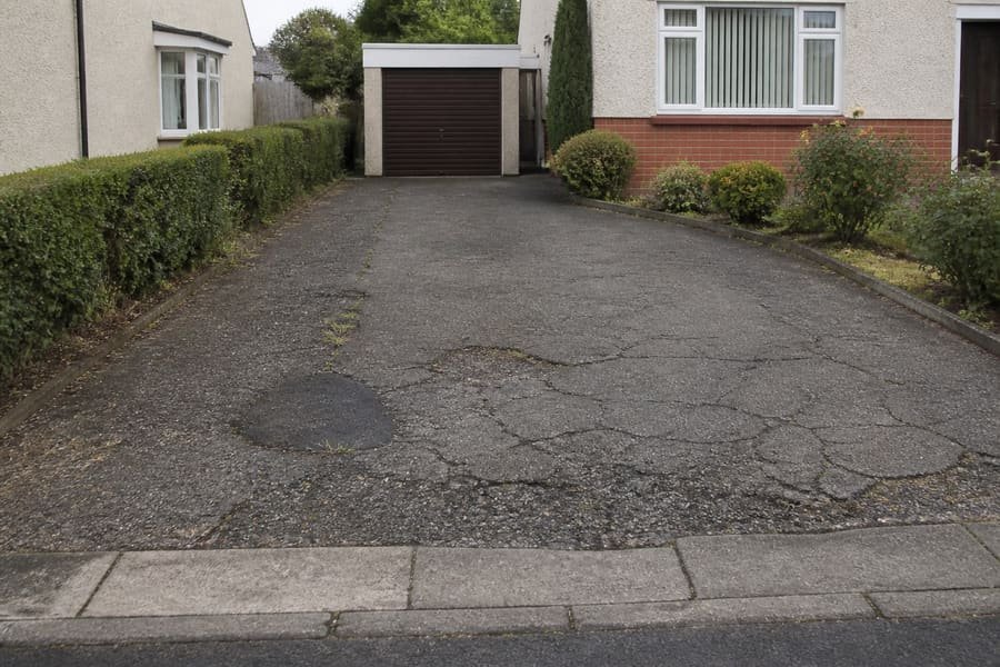 Worn driveway with cracked patched tarmac and crumbling edges.